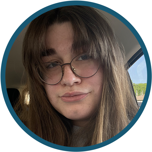Portrait of a person with long brown hair and glasses, softly lit in a car