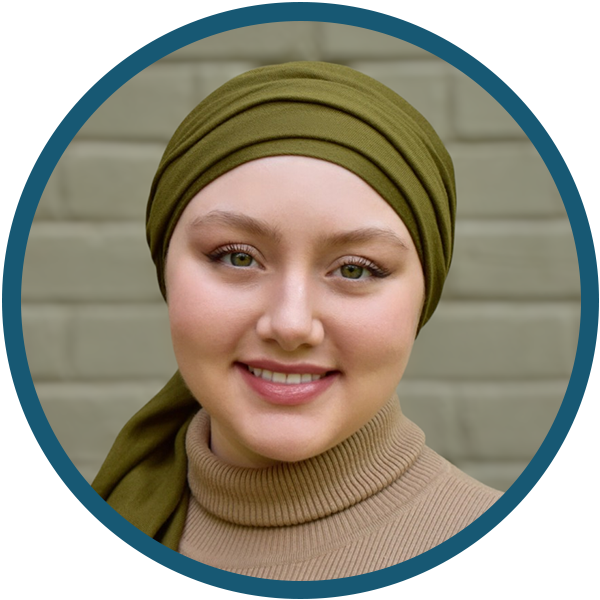Smiling person in an olive headscarf and tan sweater against a light brick wall.