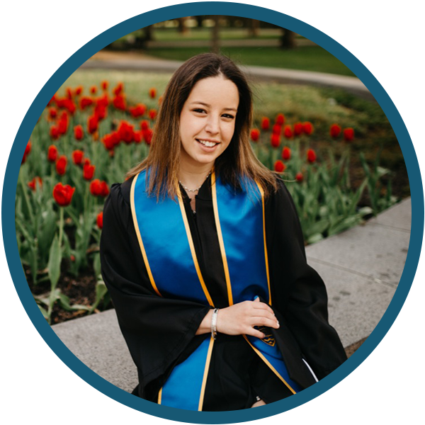 Graduate in black gown with blue stole sitting by red tulips outdoors