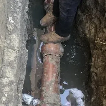 Person standing on a broken water pipe in a muddy trench.