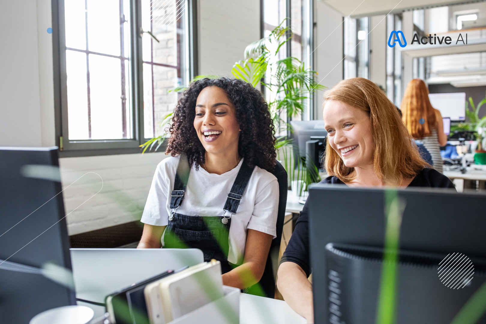 Two women smile at a computer screen in a bright office. One wears overalls, the other a black shirt.