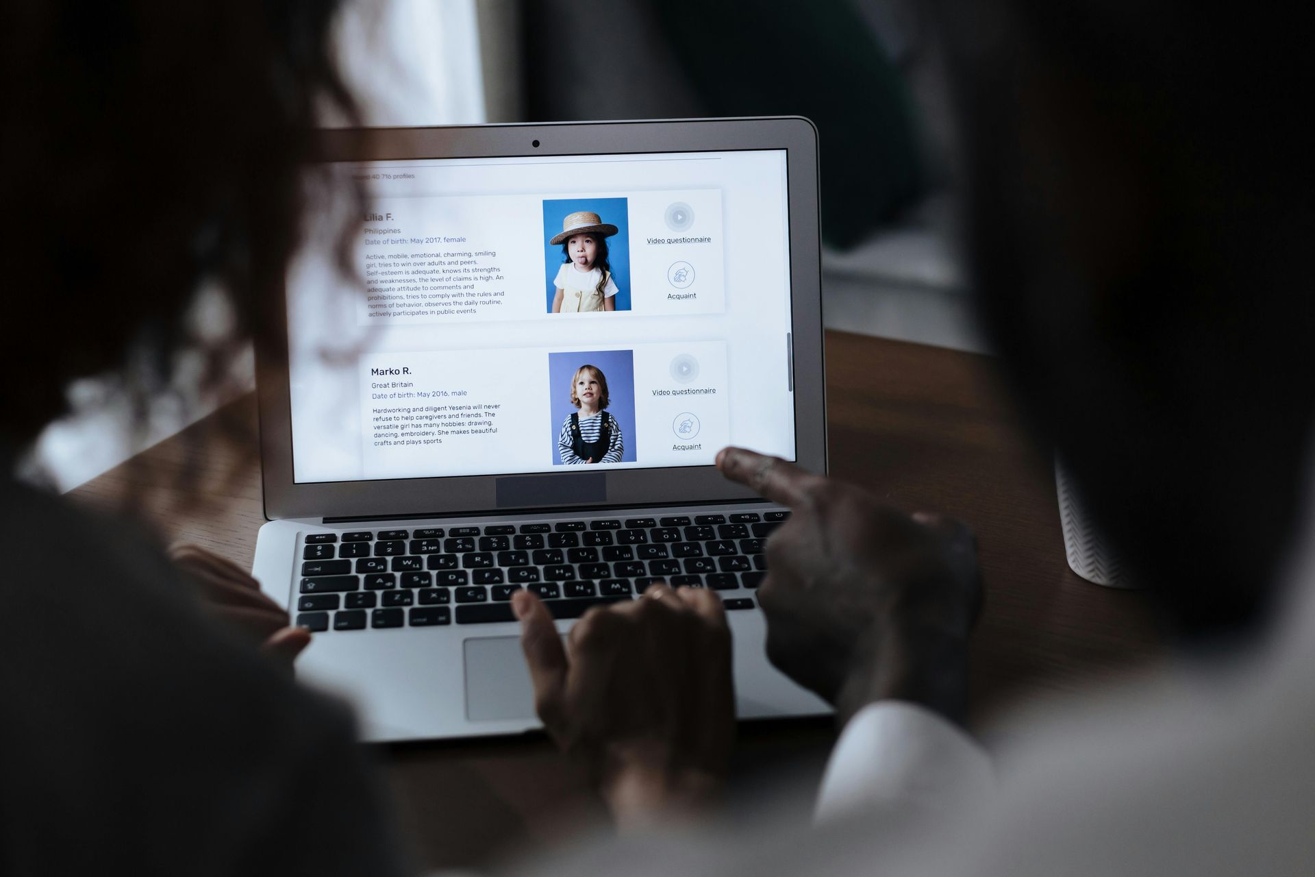 Two people looking at a laptop screen displaying profiles with children's photos. A finger points at the screen.