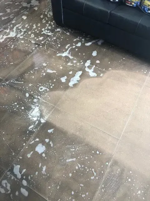 Soapy Water Spilled on Brown Tiled Floor, Near a Black Couch — Phil Lindfield's Big Red Carpet Cleaning In Taree, NSW