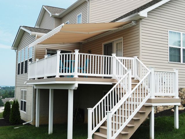 Tan retractable awning over a white-railed deck with stairs, attached to a beige two-story house.