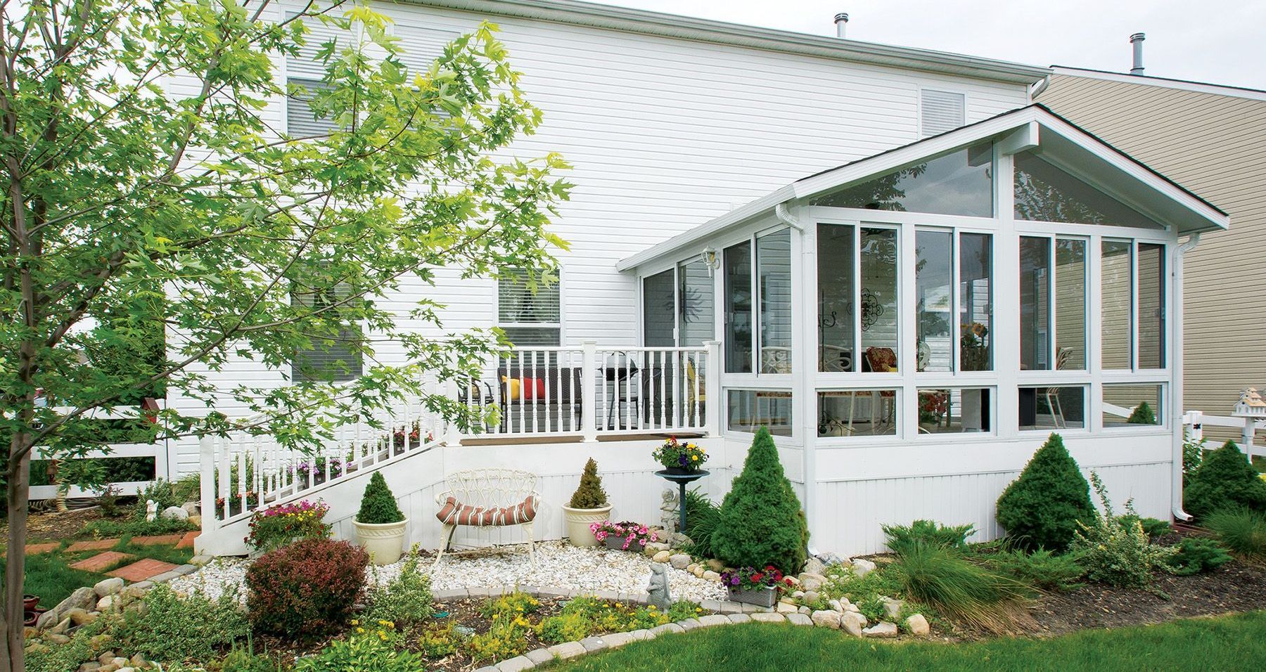 White house with a screened-in porch, deck, and garden. The porch has large windows, with a deck and white railing. The garden has shrubs and a winding path.