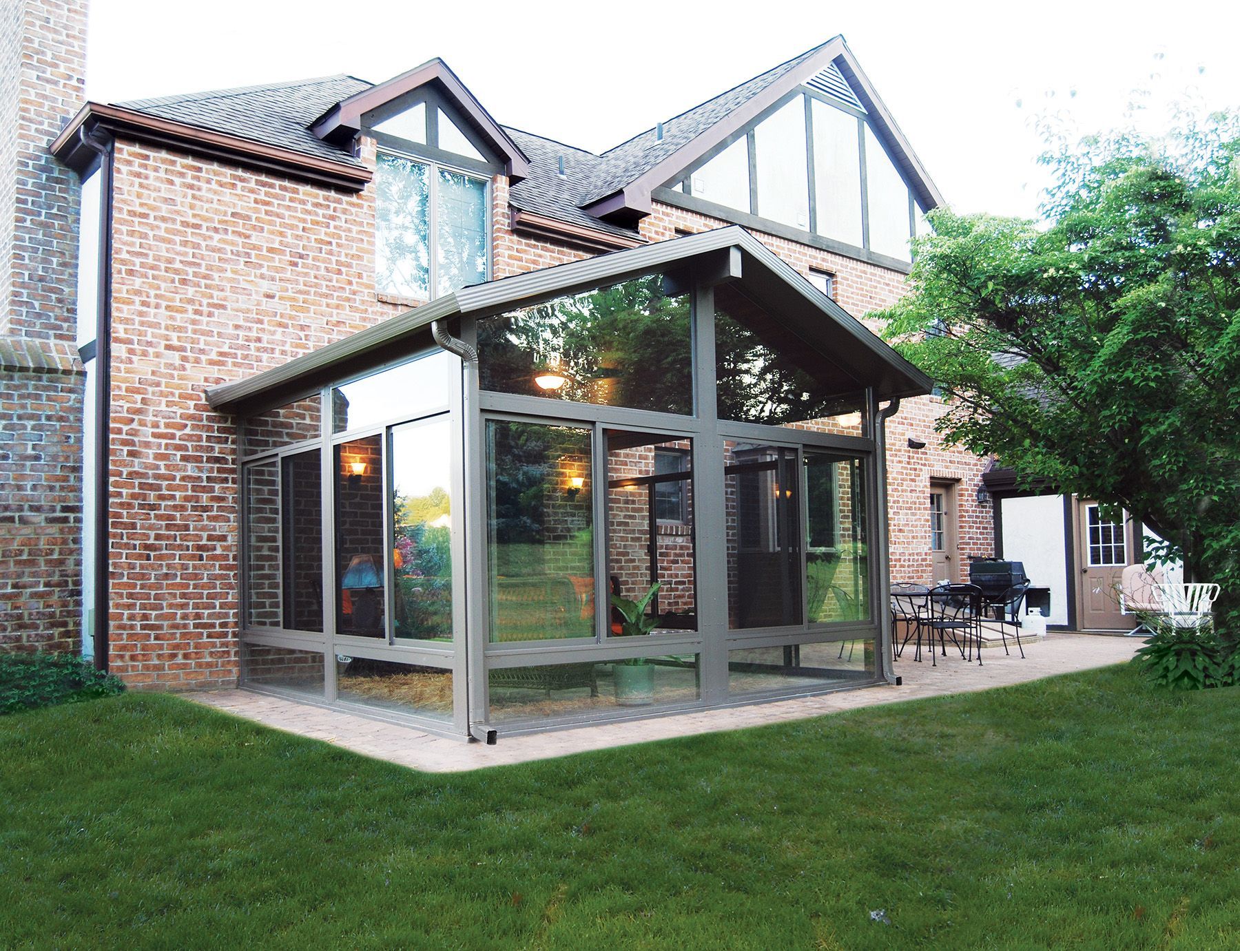 A sunroom with large glass windows and a brown frame attached to a brick house, with a patio and green lawn in the backyard.