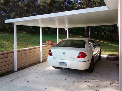 White car parked under a white carport with a brick wall and green grass background.