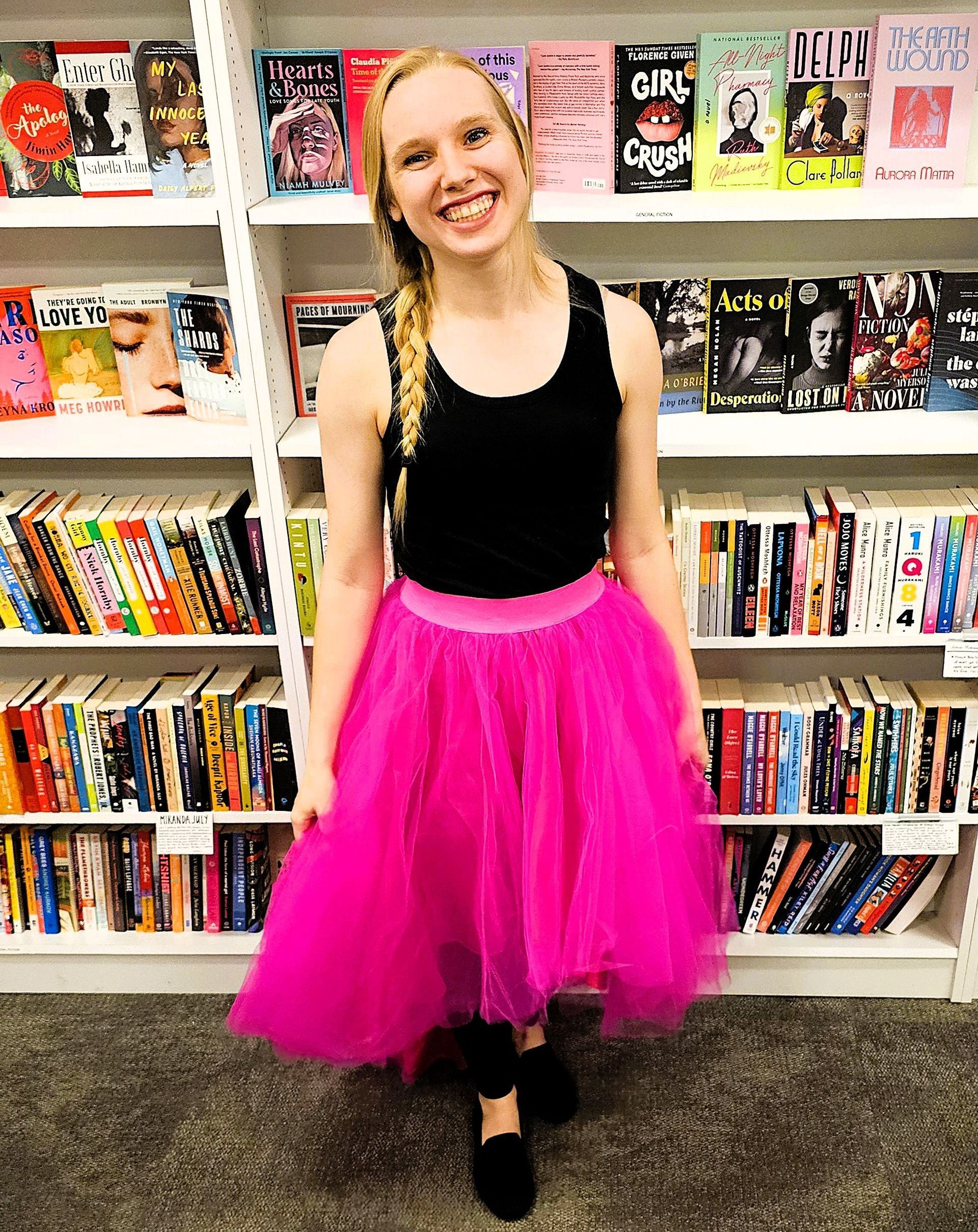 Smiling Author standing in front of library shelves wearing a black top and bright pink tulle skirt