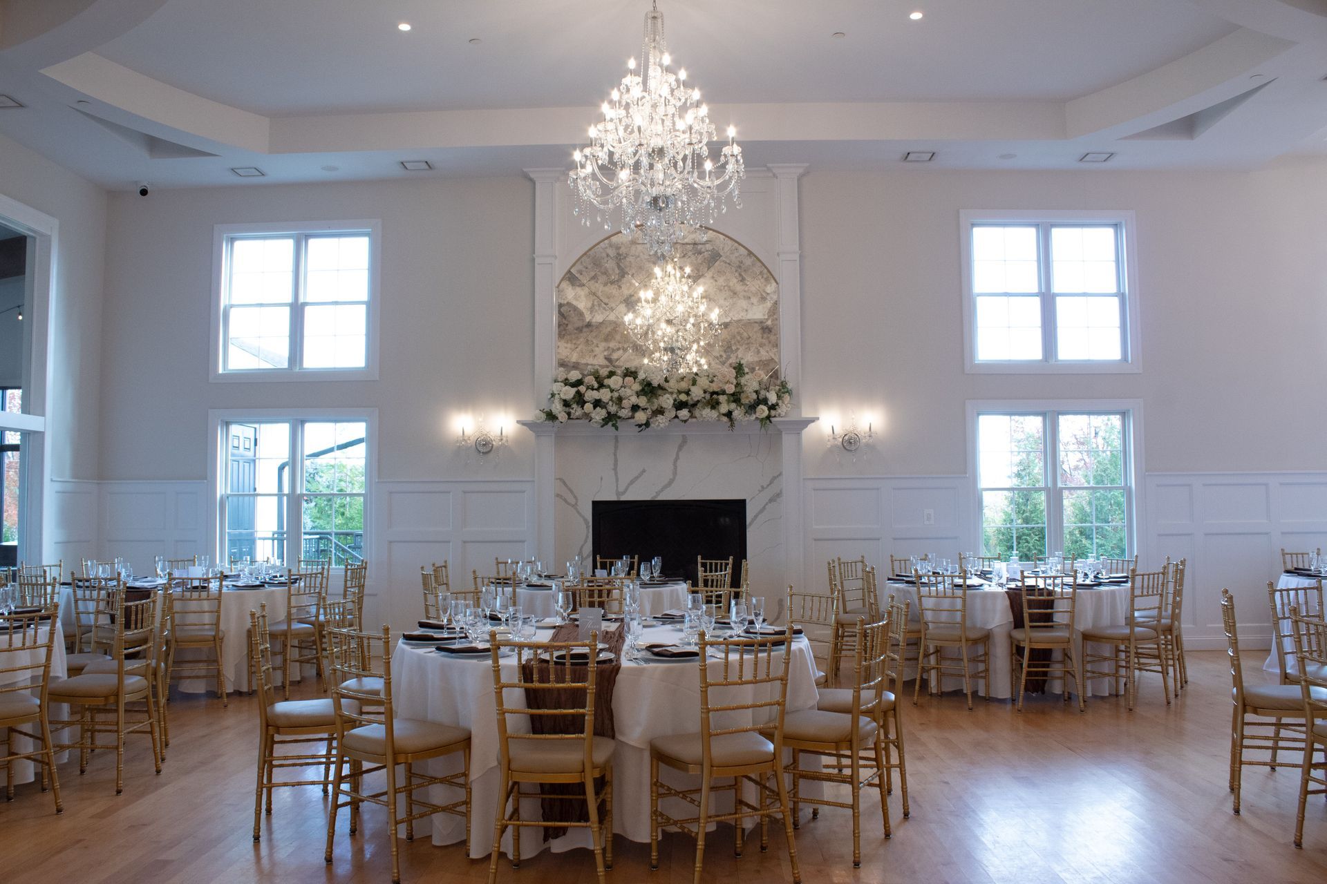 Elegant banquet hall with round tables set for a wedding reception. White walls, wood floor, chandelier.