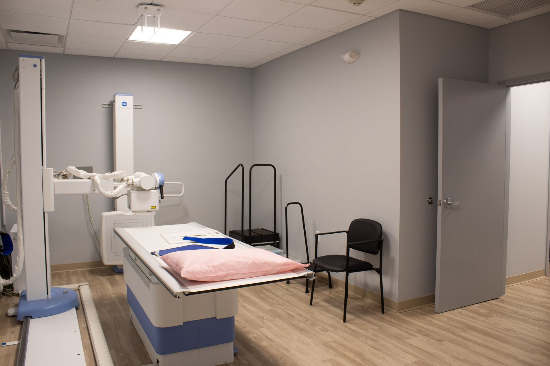 Medical imaging room with an X-ray machine, examination table, chair, and open doorway.