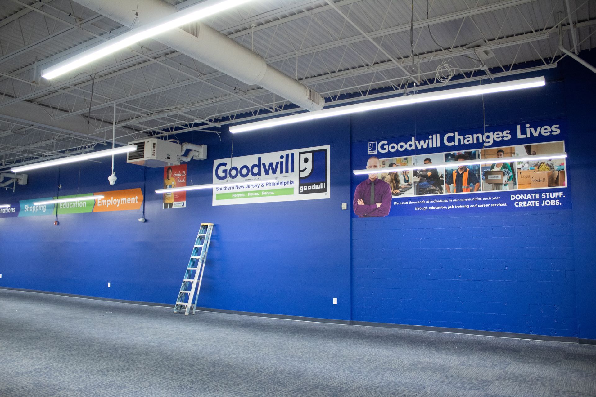 Blue wall with Goodwill signage and a ladder. Fluorescent lights and a gray carpeted floor are also visible.