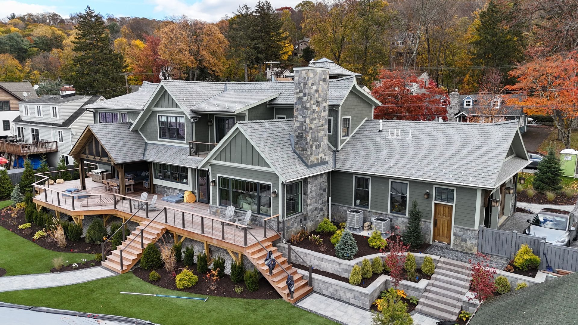 Large green house with stone chimney, wooden deck, and landscaping. Autumn foliage in background.