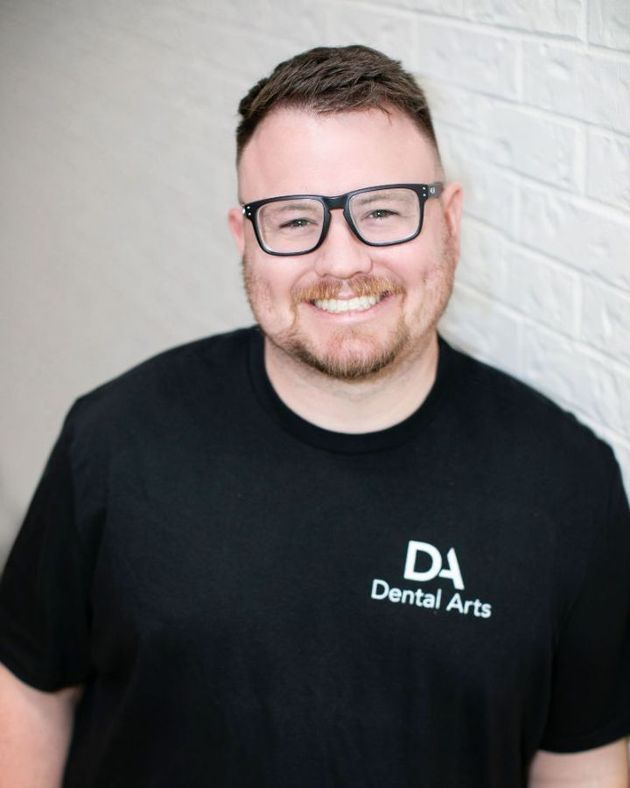 A man wearing glasses and a black shirt is standing in front of a white brick wall.