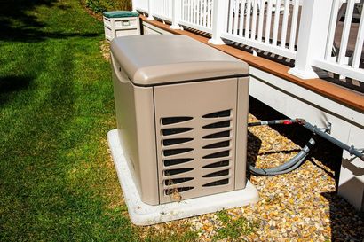 Tan home generator on a concrete pad next to a white porch, green grass.