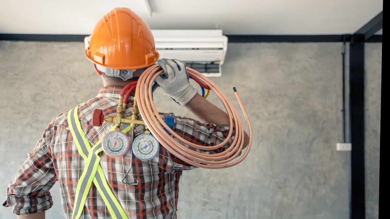 HVAC technician carrying copper tubing, wearing a hard hat and safety vest, near an AC unit.
