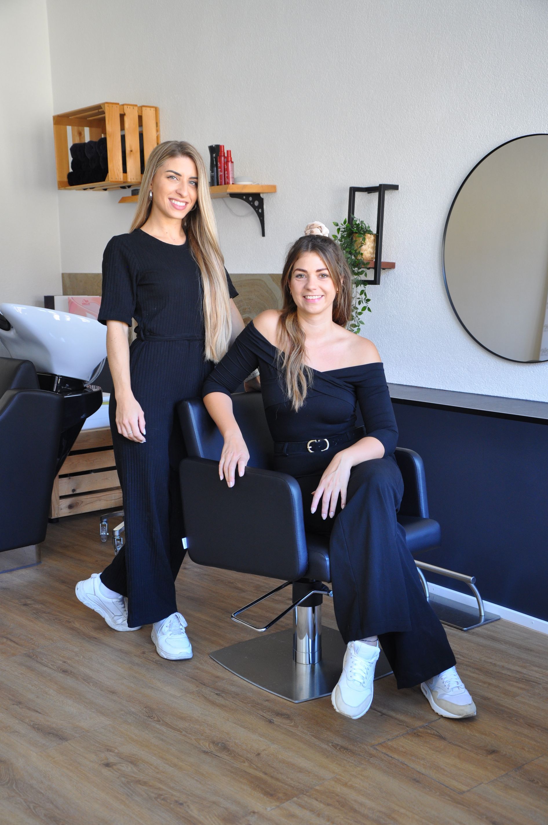 Zwei Frauen stehen nebeneinander in einem Friseursalon. Eine der Frauen sitzt auf einem Stuhl.