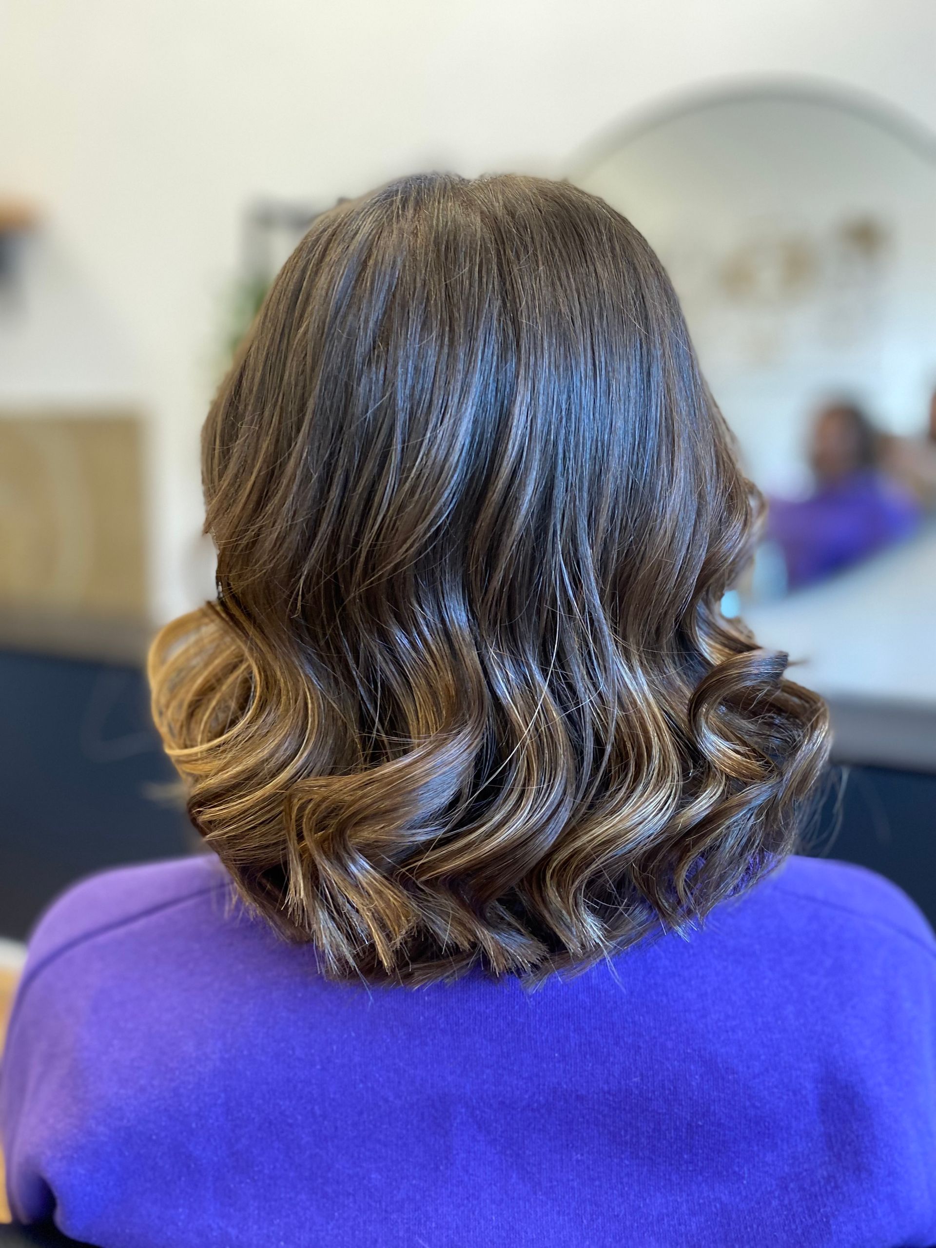 Eine Frau in einem lila Hemd sitzt auf einem Stuhl und hat ihr Haar zu einem Pferdeschwanz gebunden.