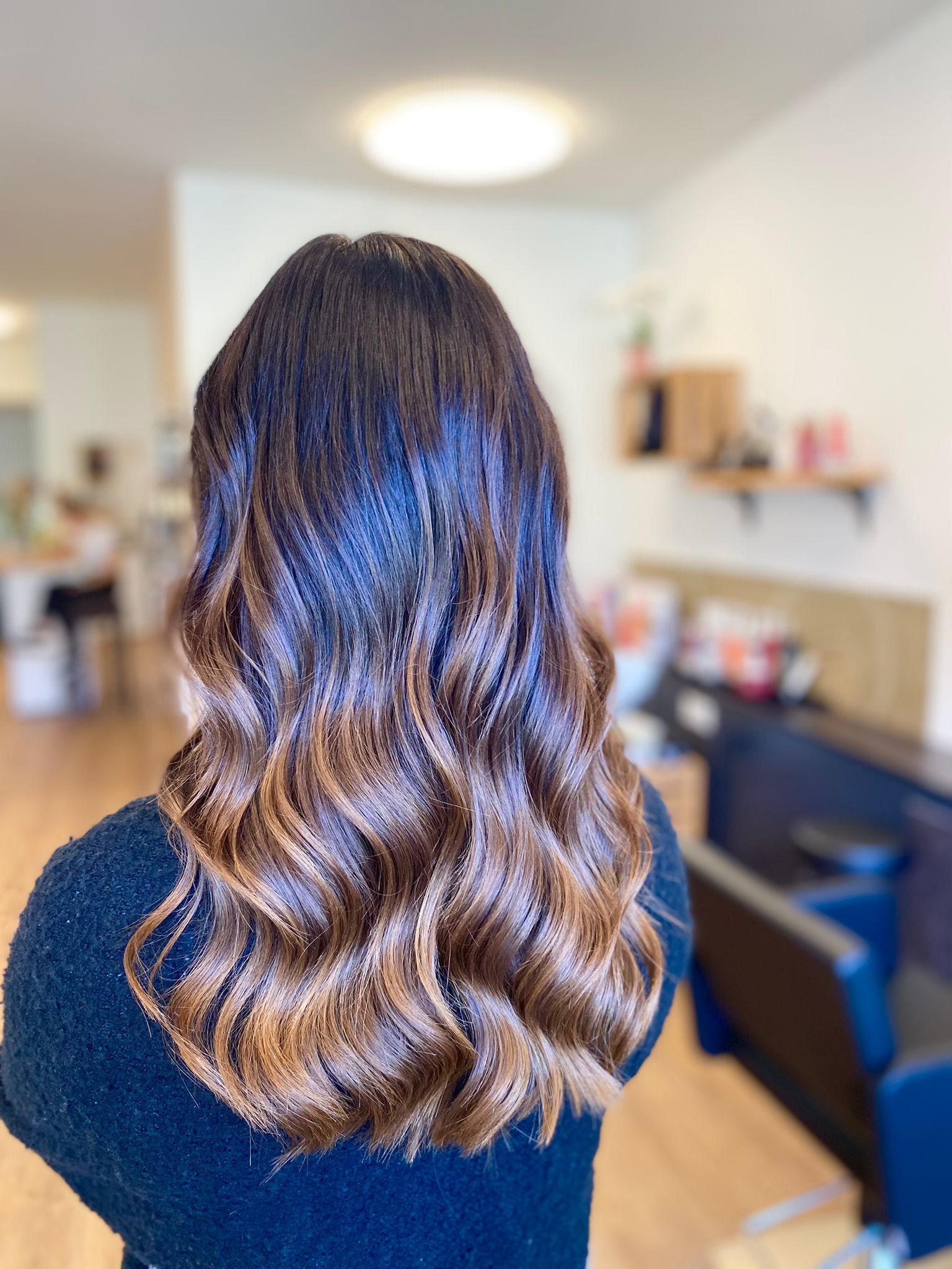 Eine Frau mit langen braunen Haaren sitzt in einem Salon auf einem Stuhl.