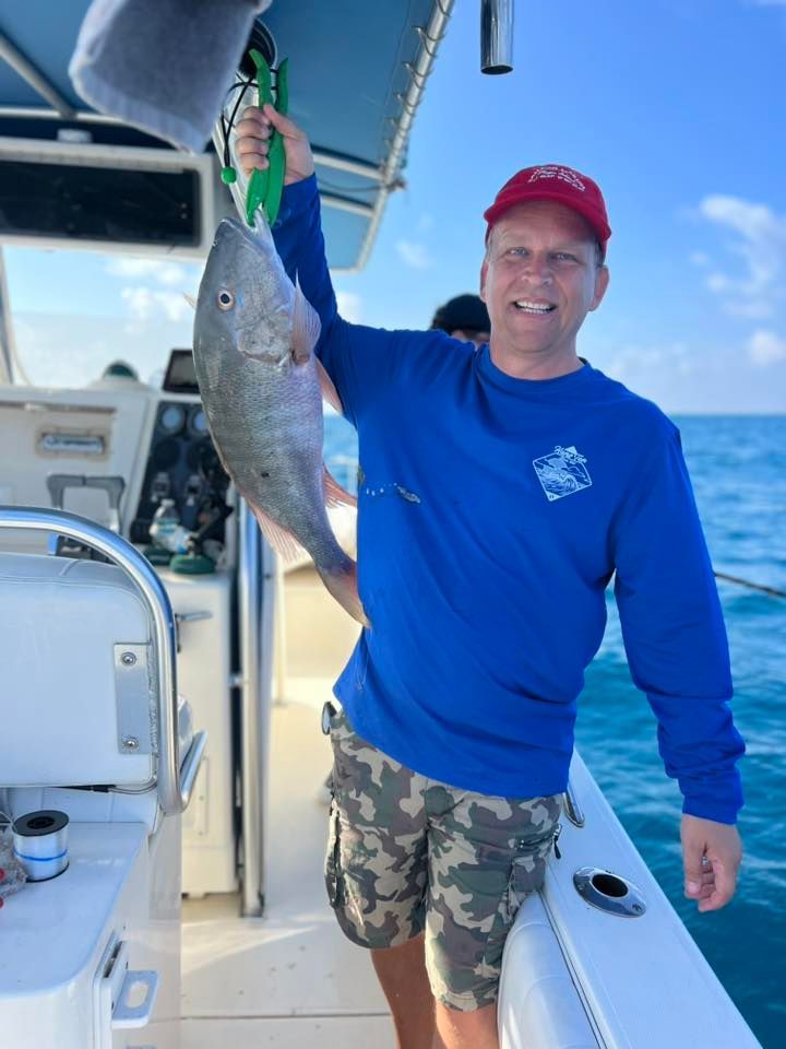 Man on a boat, holding up a fish. Blue shirt, camo shorts, red cap. Smiling. Ocean background.
