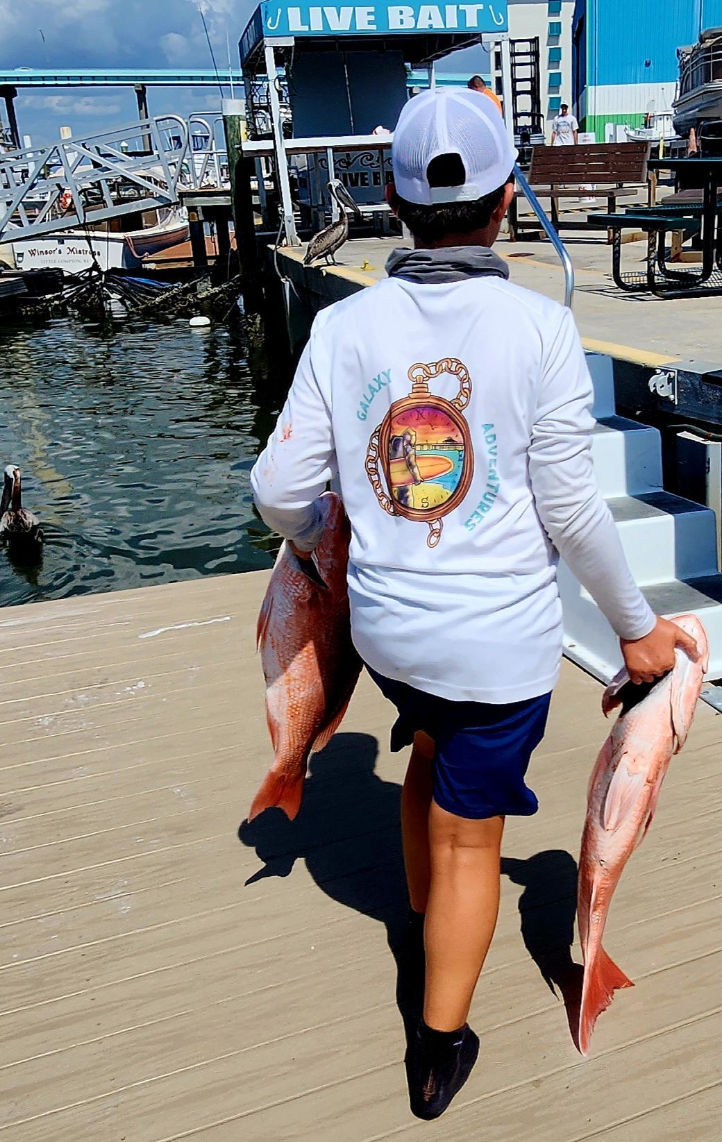 Person carrying two large red fish on a dock.  Blue shorts, white long-sleeve shirt, and a white cap.  Sunny day.