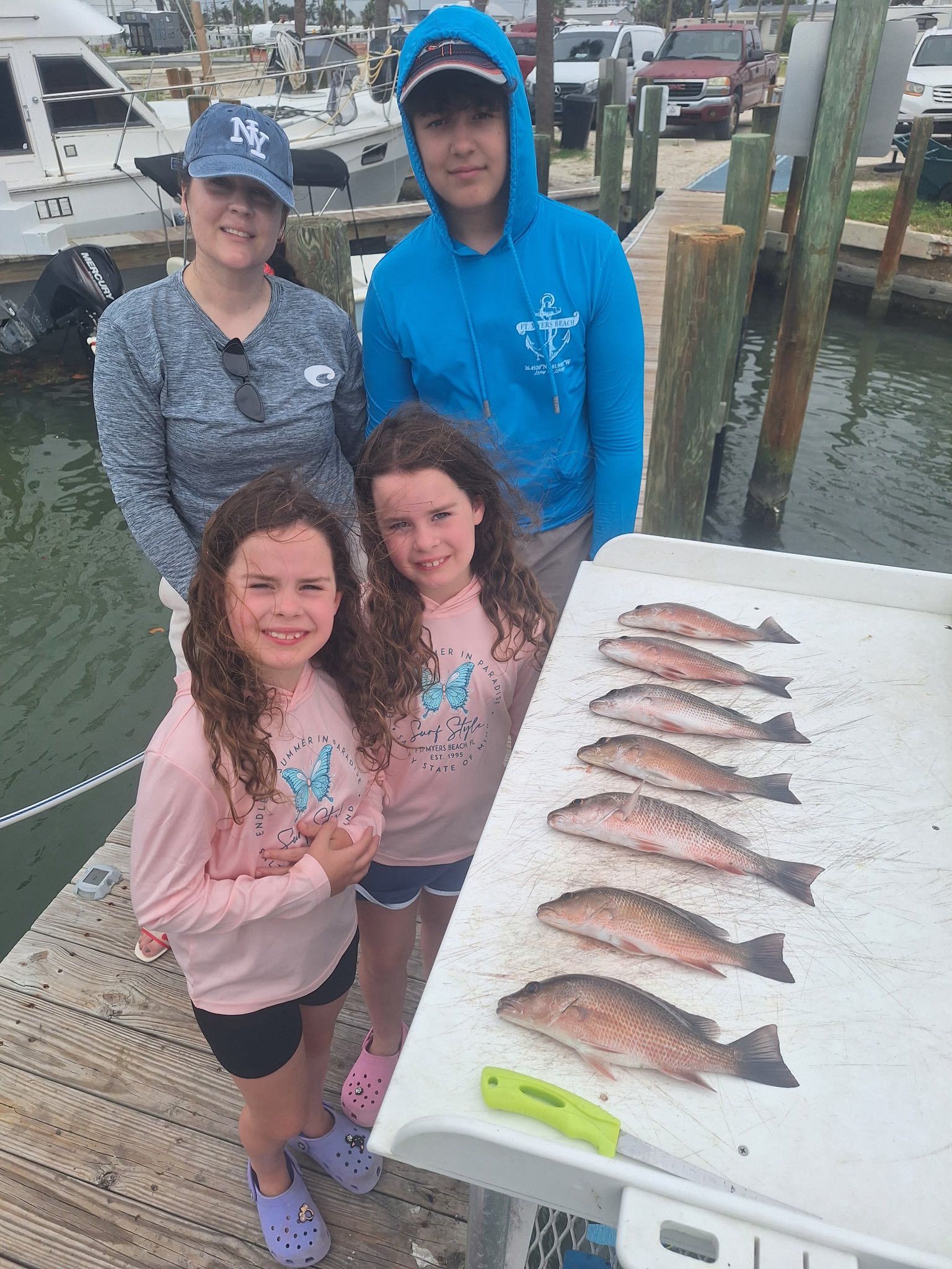Family posing with a fish catch at a dock. Two girls smile, adults behind. Fish laid out on a table.