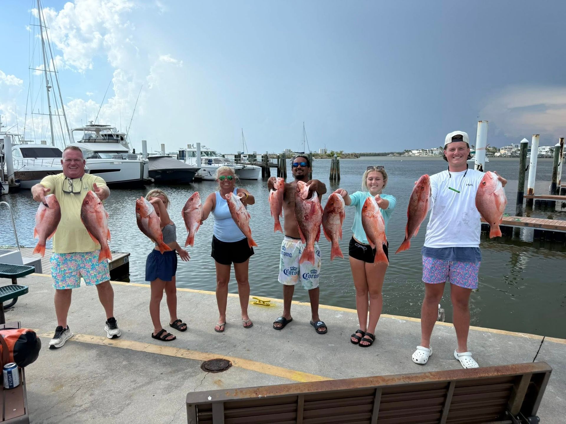 Group holding red snapper, smiling, at a dock with boats, sunny day.