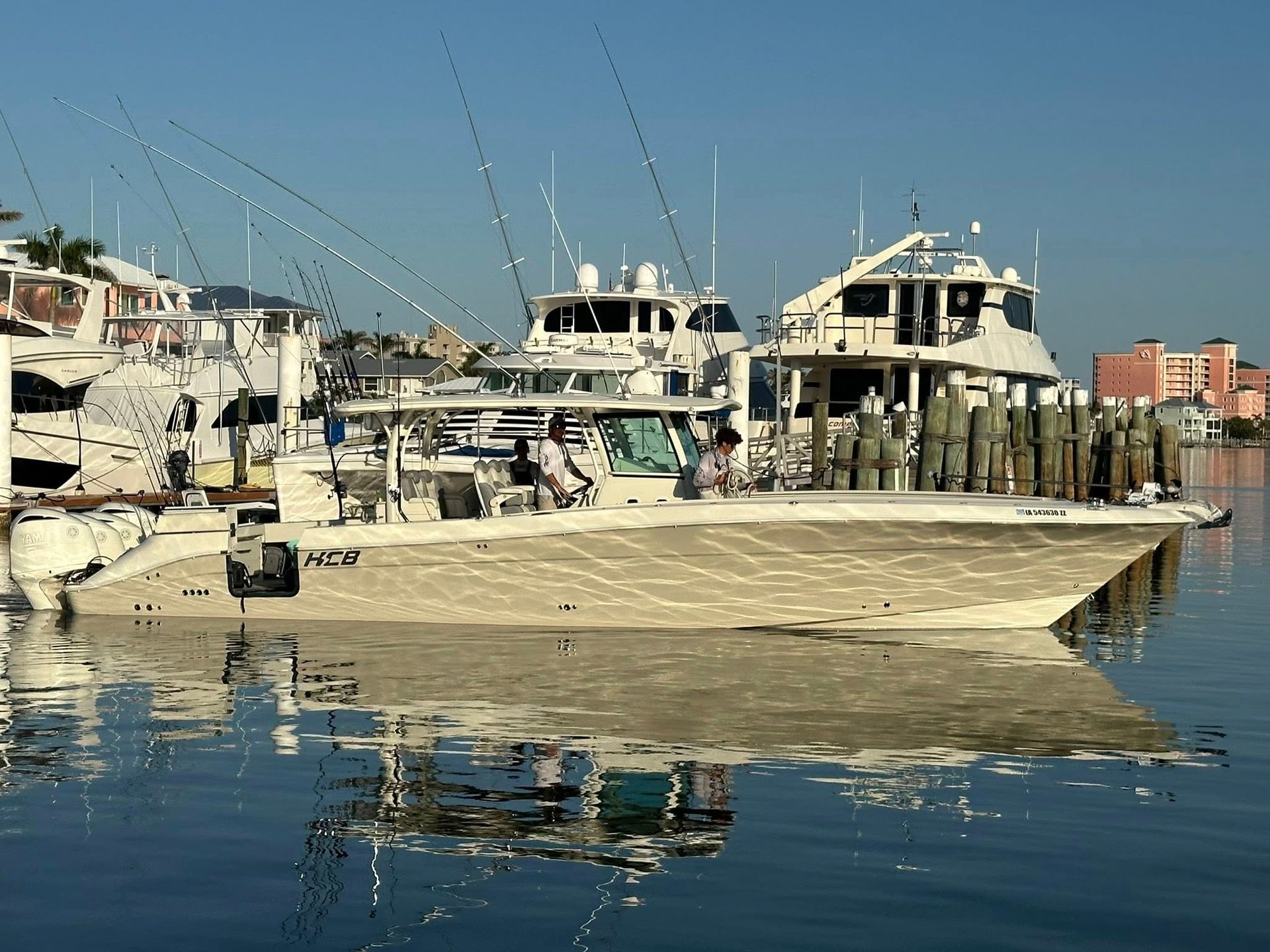 Boats docked at a harbor, reflections in the water. Clear sky, buildings in background.