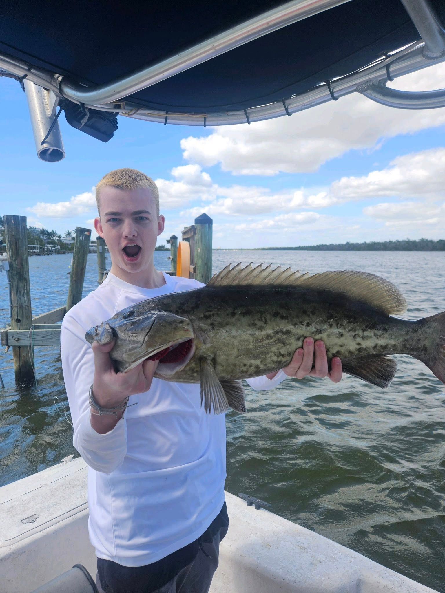 Teenager on a boat holding a large, dark fish, mouth open. Outdoors, sunny, water in background.