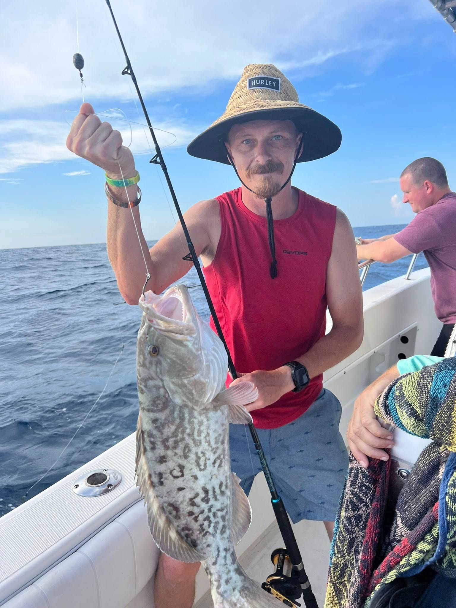 Man on a boat holding a spotted fish he caught. He wears a hat and red shirt, with another man behind him. Ocean in background.