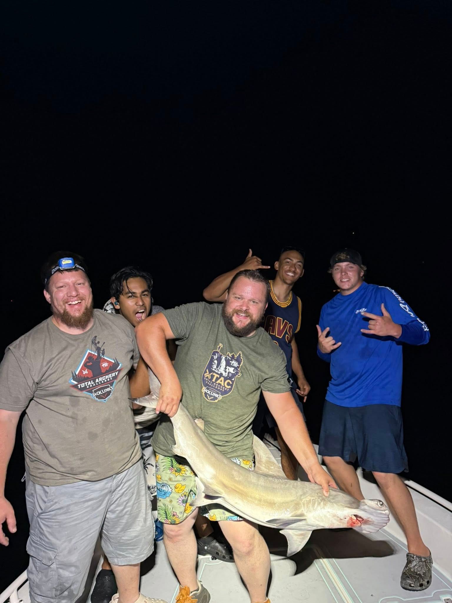 Five people on a boat at night hold up a large fish, all smiling.