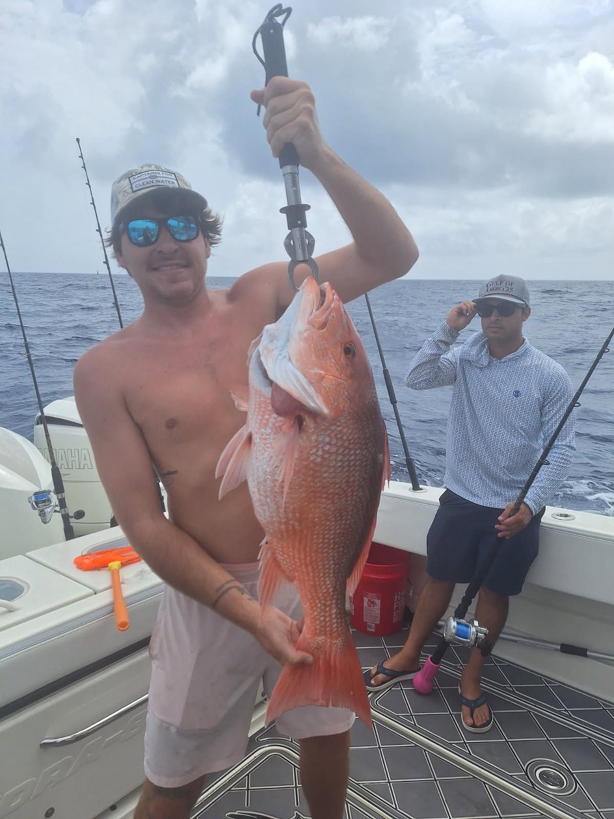 Man on a boat holds up a large red snapper fish, smiling. Another man stands behind him. Ocean background.