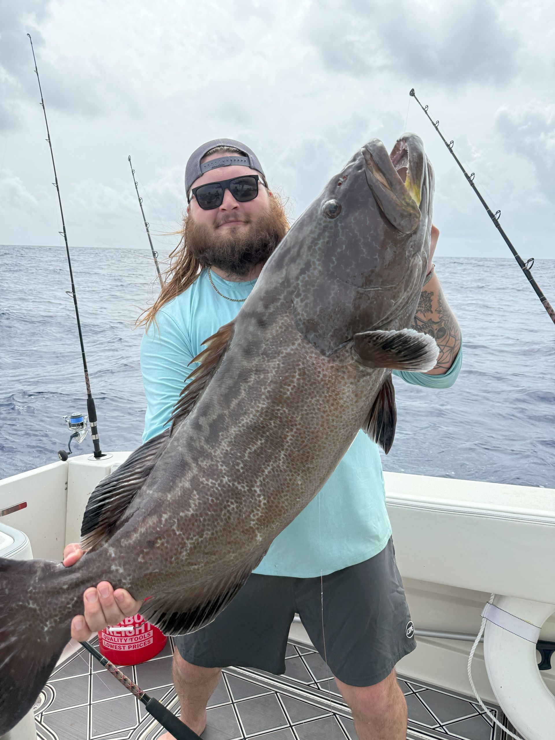 Man on a boat holding up a large, dark-colored fish with open mouth, ocean background.
