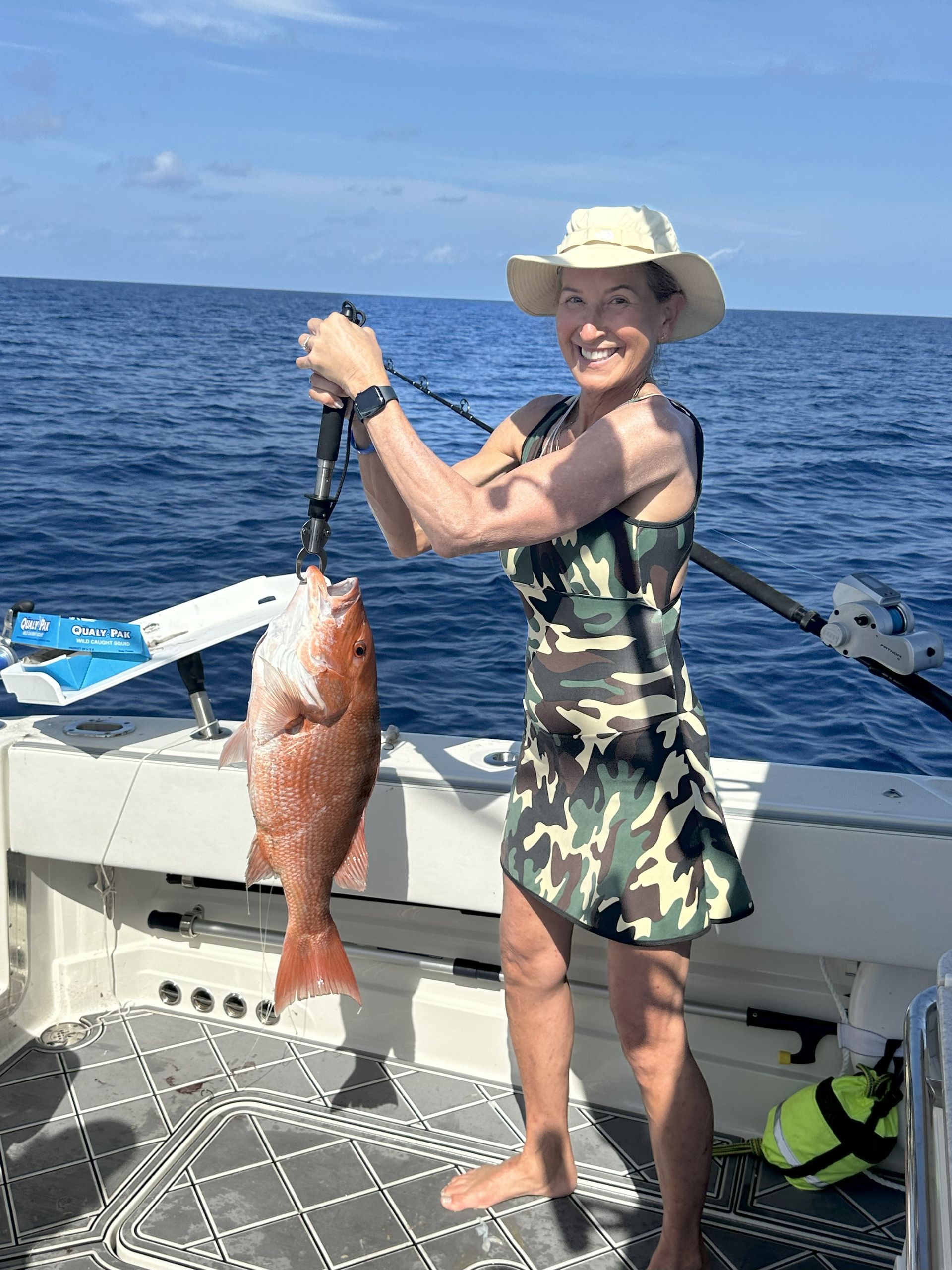 Woman on a boat holding up a large red fish, smiling. Wearing a camo dress and sun hat.
