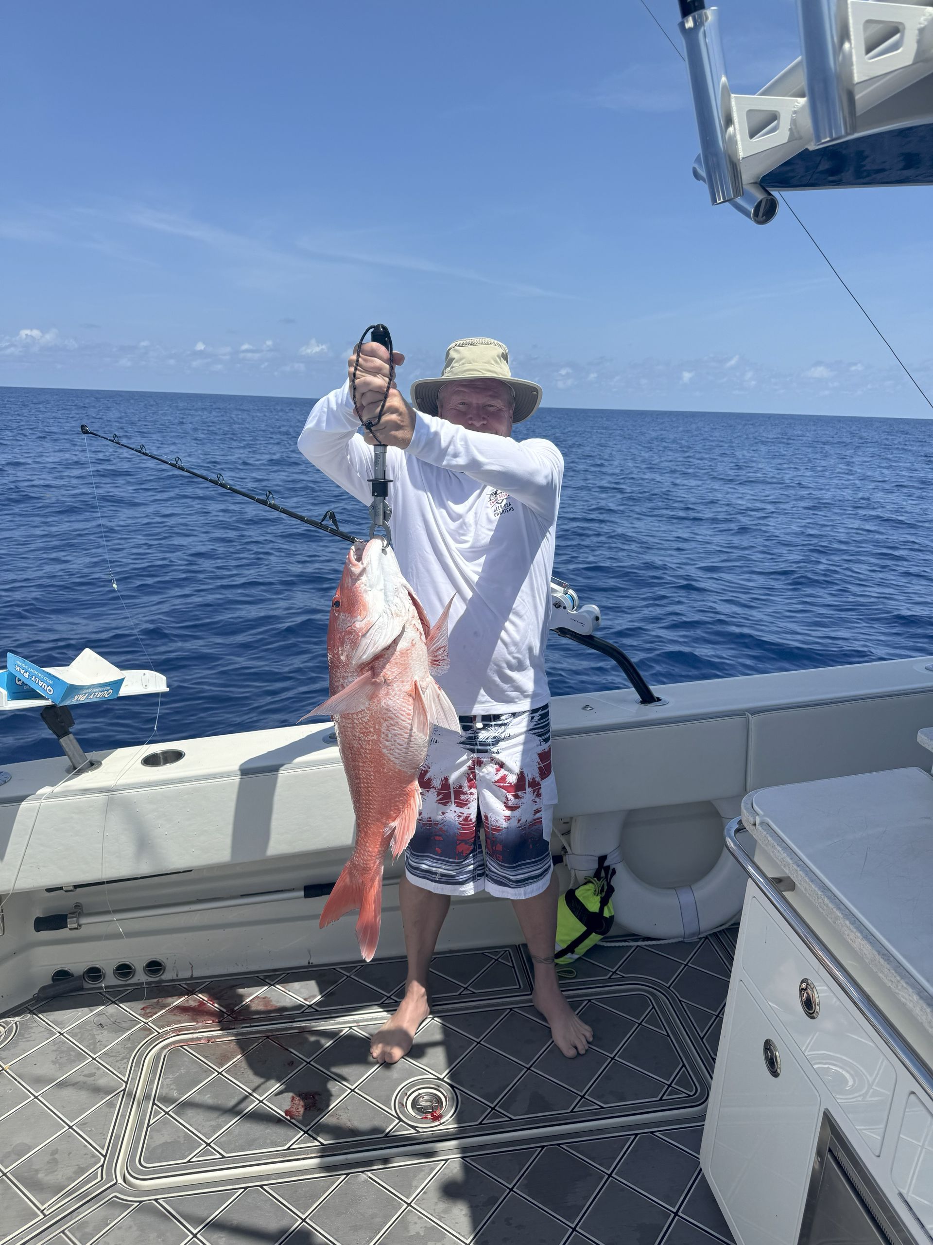 Man on boat holding up a large, red fish. Blue water, sunny sky.