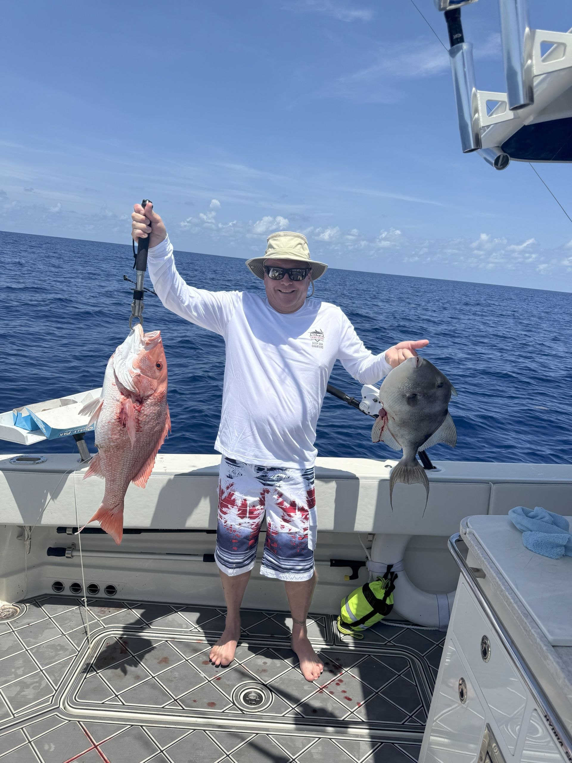 Man on boat holding up a red snapper and a gray triggerfish. Blue sea, sunny day.