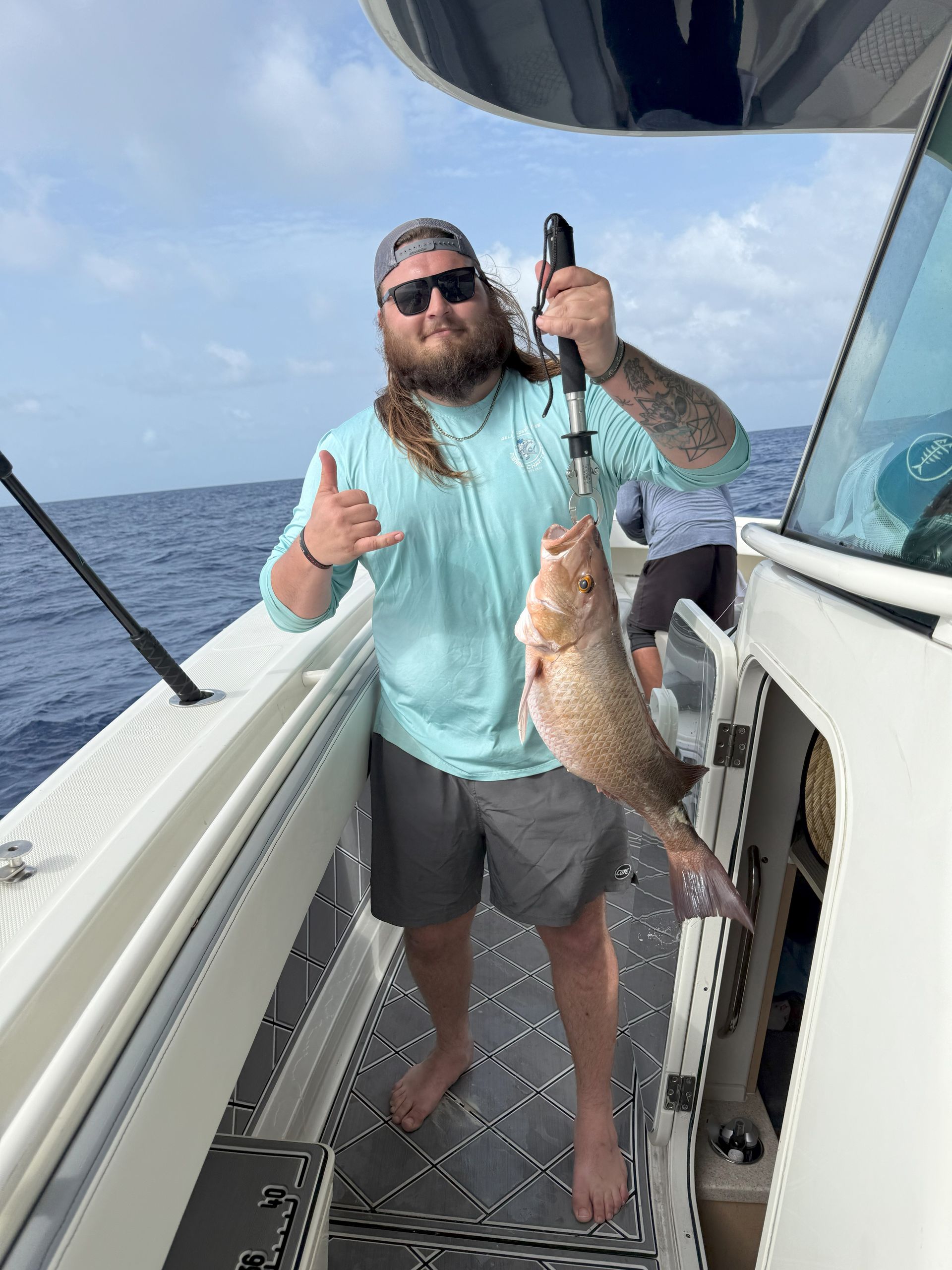 Man on a boat fishing, holding up a grouper fish, giving a shaka sign.