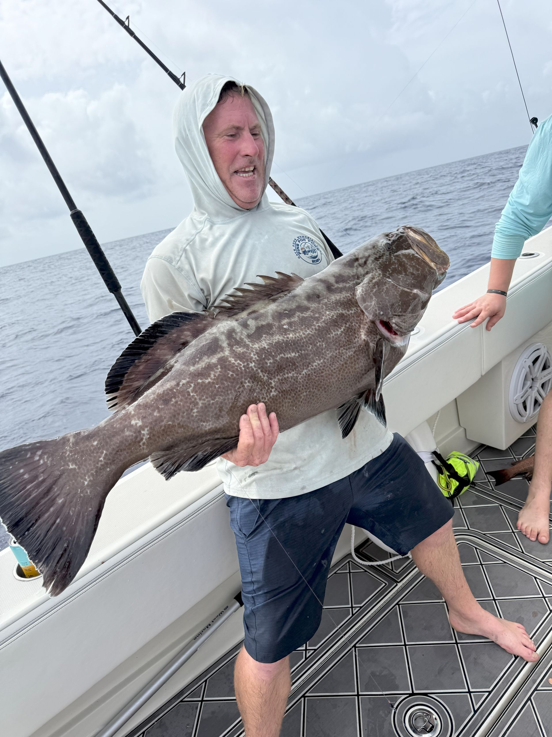 Man on boat holding a large, spotted grouper. He's wearing a hooded jacket and shorts. Ocean in the background.