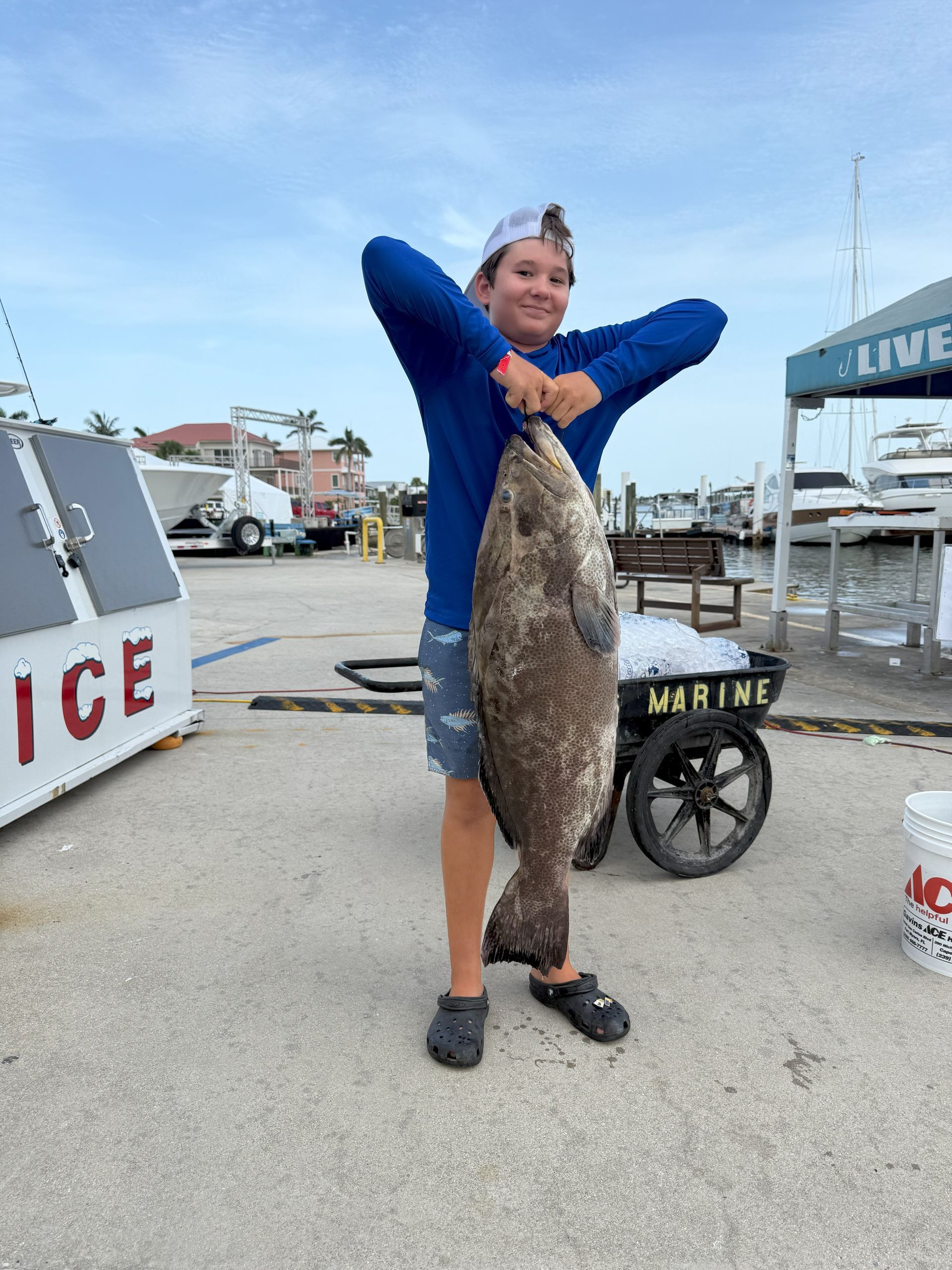Boy holding a large, dark fish on a dock, smiling. Blue sky, ice cart, boats in the background.