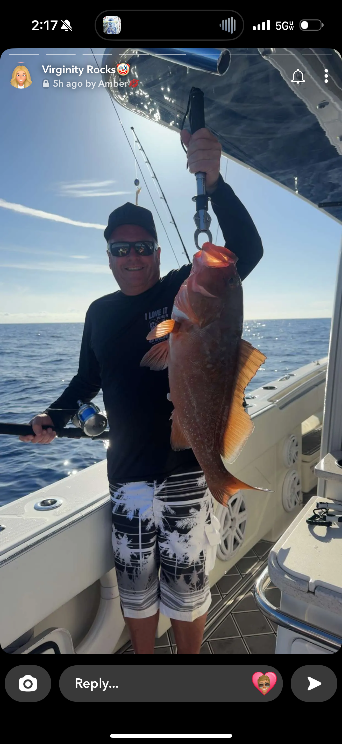 Man on a boat holds up a large fish he caught. He wears a black shirt and sunglasses, against a blue sea.