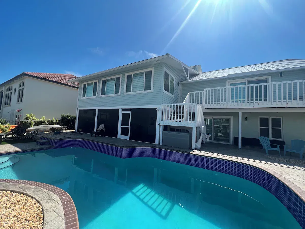 Backyard with a pool and two-story blue house. Deck with white railing. Sunny, blue sky.