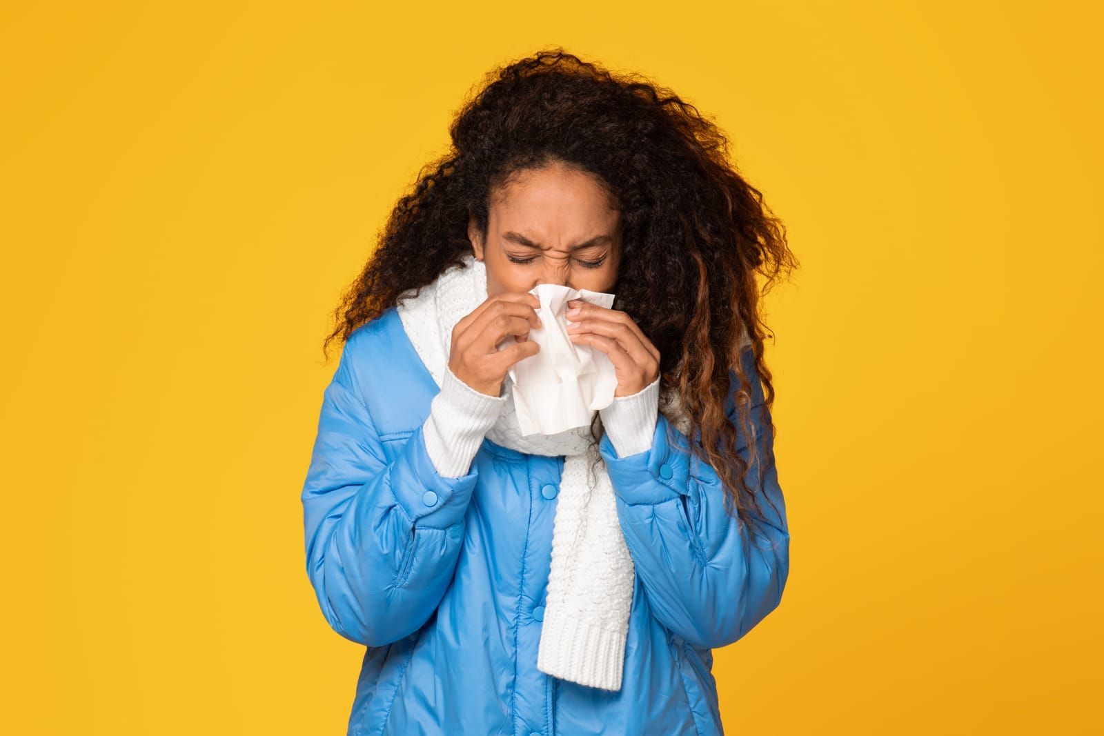 Woman wearing a blue jacket and scarf blows her nose with a tissue, eyes closed, against a yellow background.