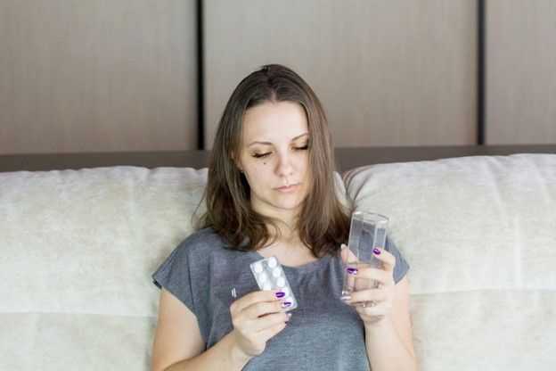 A woman is sitting on a couch holding a glass of water and a pill.