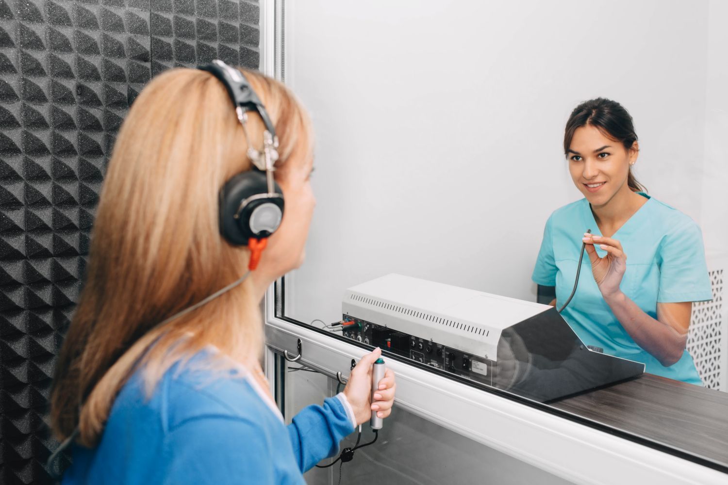 Woman in soundproof booth taking hearing test. Audiologist in blue scrubs observes.