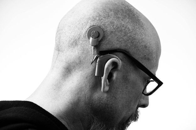 A black and white photo of a bald man wearing glasses and hearing aid