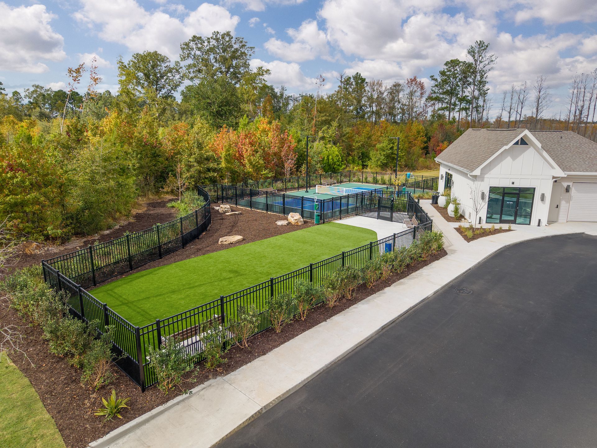 Fenced dog park with artificial turf, trees, and a white building. Asphalt road in front.