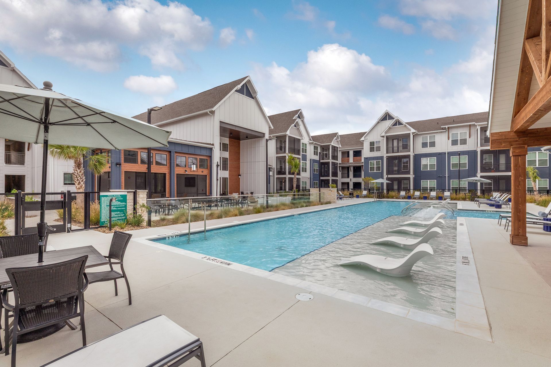 Apartment pool area with lounge chairs, tables with umbrellas, and multi-story buildings.