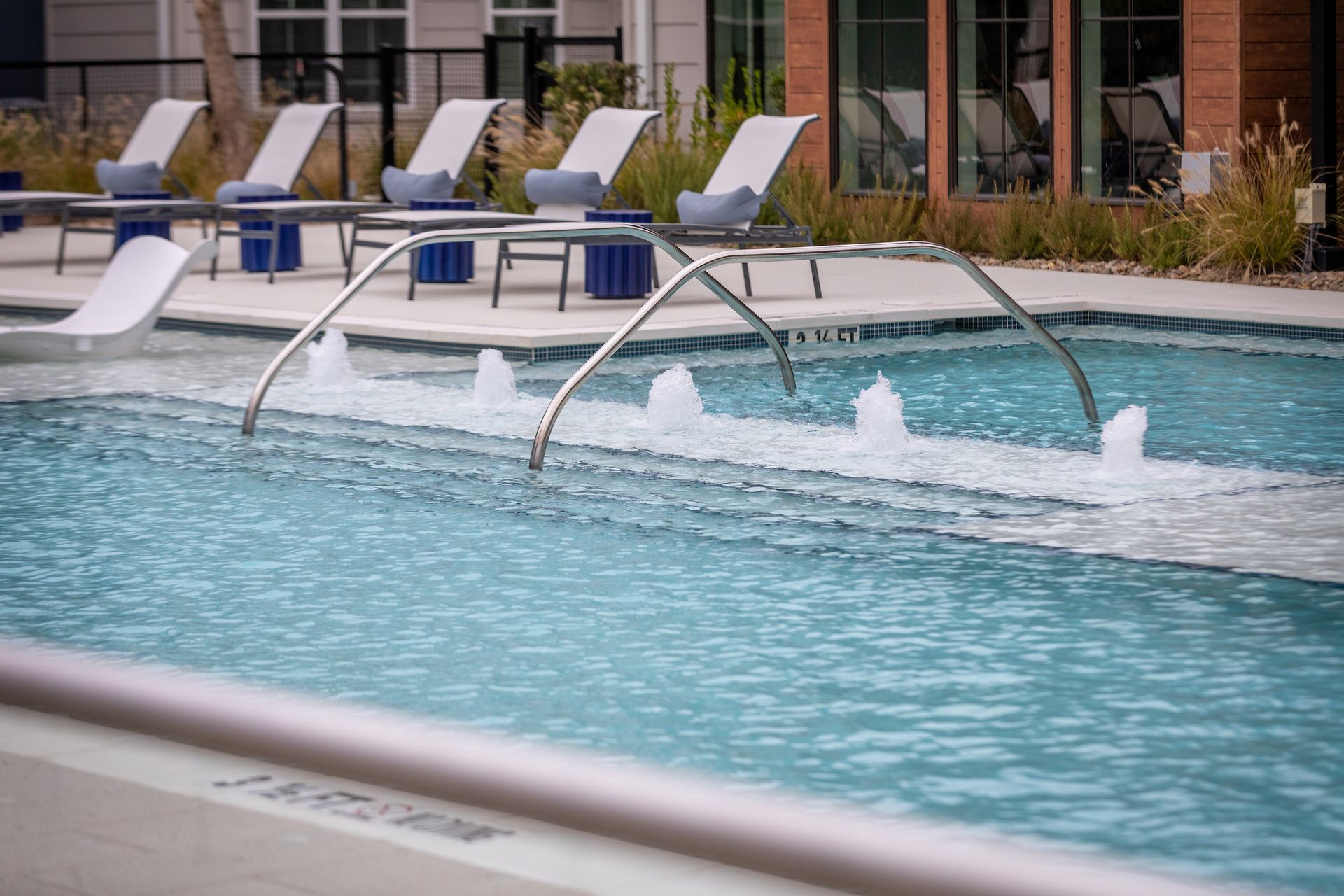 Pool with fountains, lounge chairs, and building in the background. Blue water, stainless steel rails.