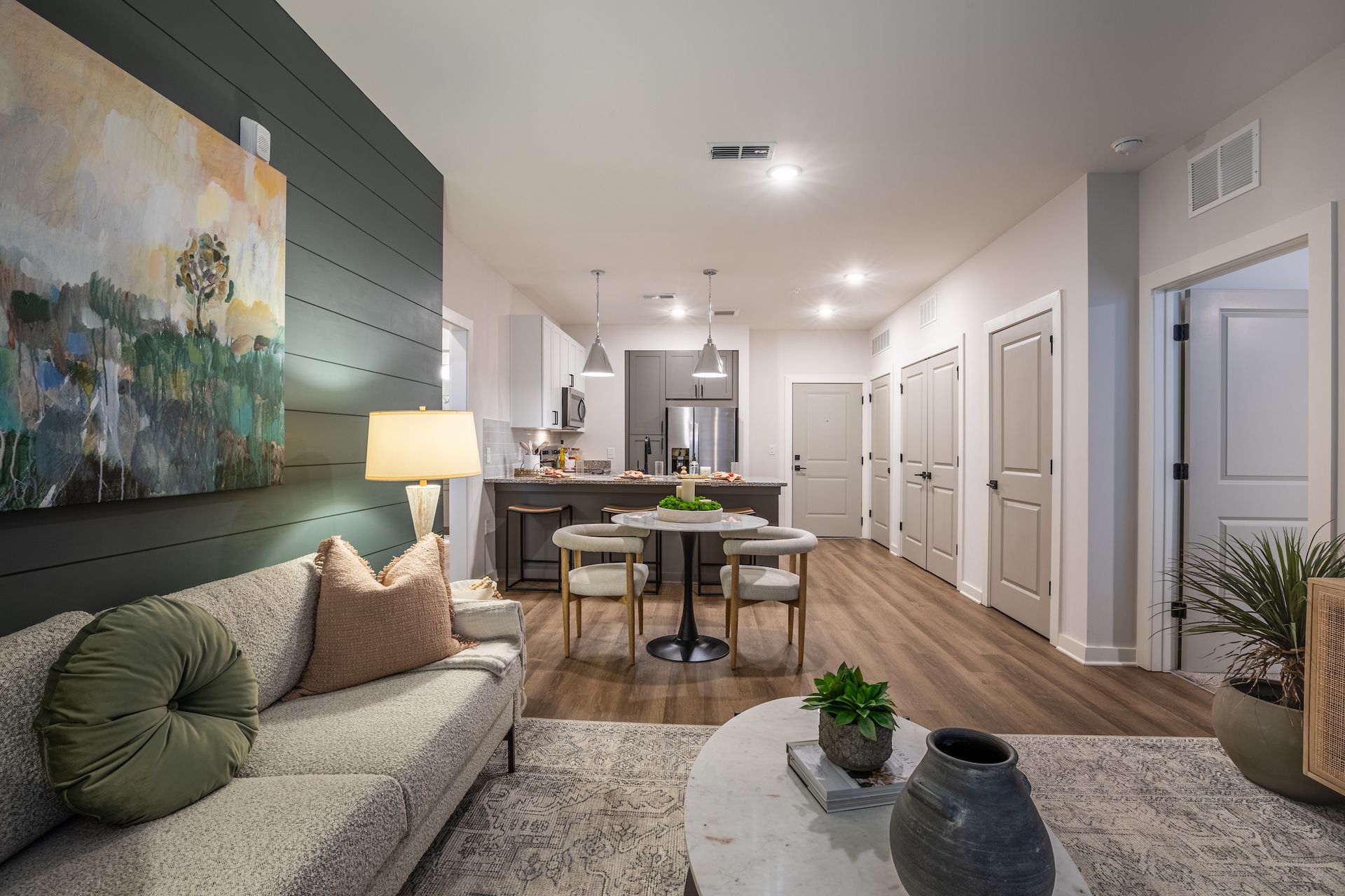 Living room with wood floors, open to a kitchen. Couch with pillows, dining table, and artwork on wall.