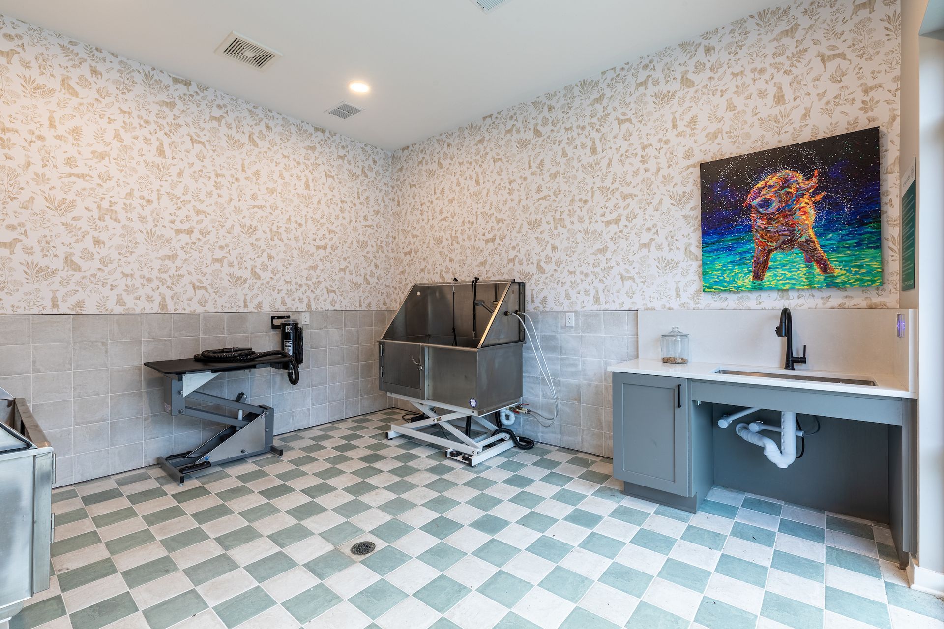 Dog grooming room with a tub, sink, and grooming table. Light gray and green tiled floor with a floral pattern wallpaper.