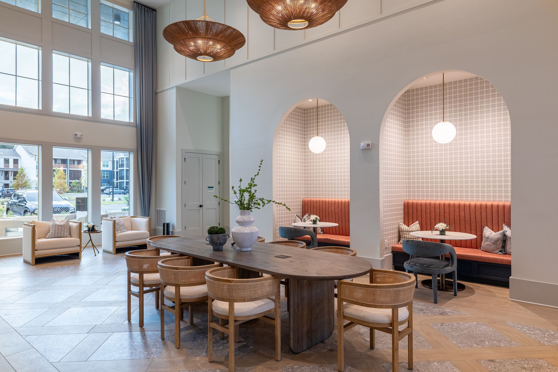 Spacious lobby with long table and chairs, round booth seating, and large windows. Beige, wood, and orange tones.